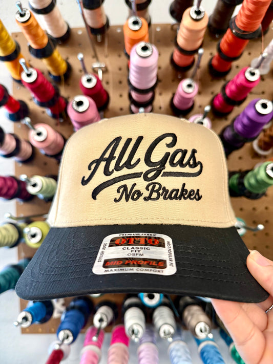 A tan and black snapback cap held by a hand in front of a pegboard filled with colorful spools of thread. The cap features black embroidered text reading 'All Gas No Brakes' on the front panel.