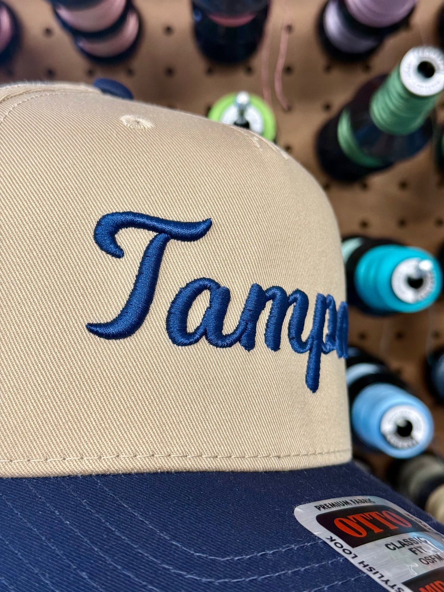 A close-up of a beige and navy blue snapback cap with 'Tampa' embroidered in blue script on the front, set against a background of colorful spools of thread on a pegboard.